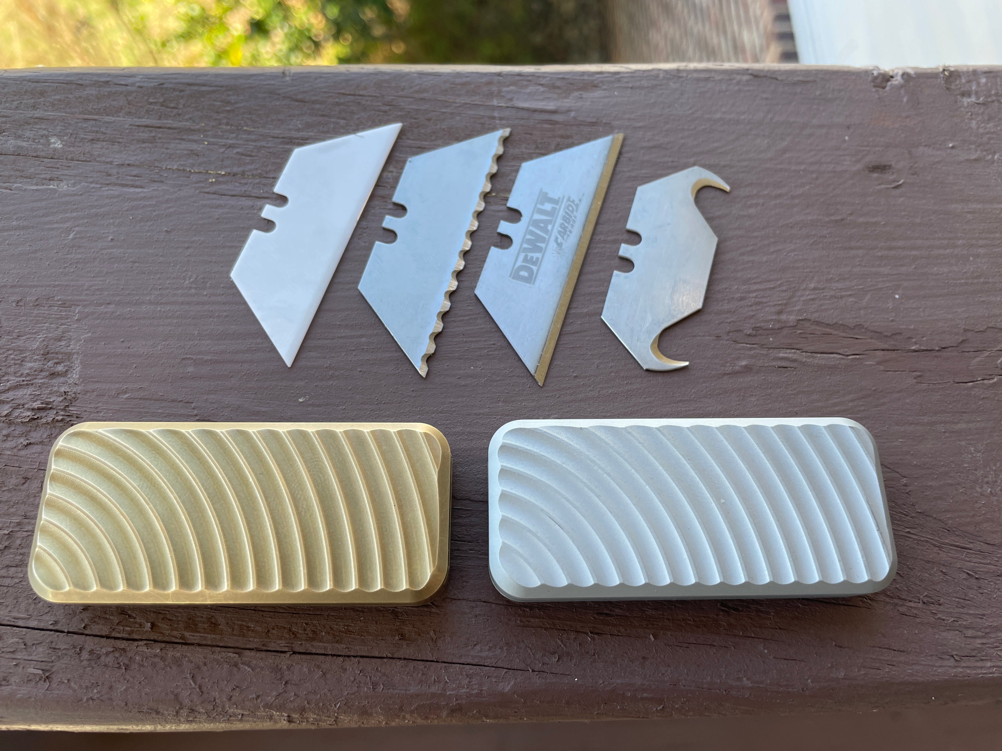 Assortment of utility knife blades and handles on a wooden surface, including DEWALT carbide blades and two textured Zero Utility Knives in gold and silver.