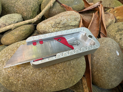 Gray Zero utility knife by RiveryMFG with open lid, red blade holder and spring on rocks.