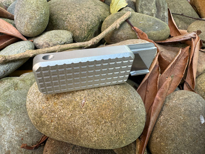 Gray fidget knife Zero by riverymfg side view with grenade-textured handle, lanyard hole and open lid resting on stones.